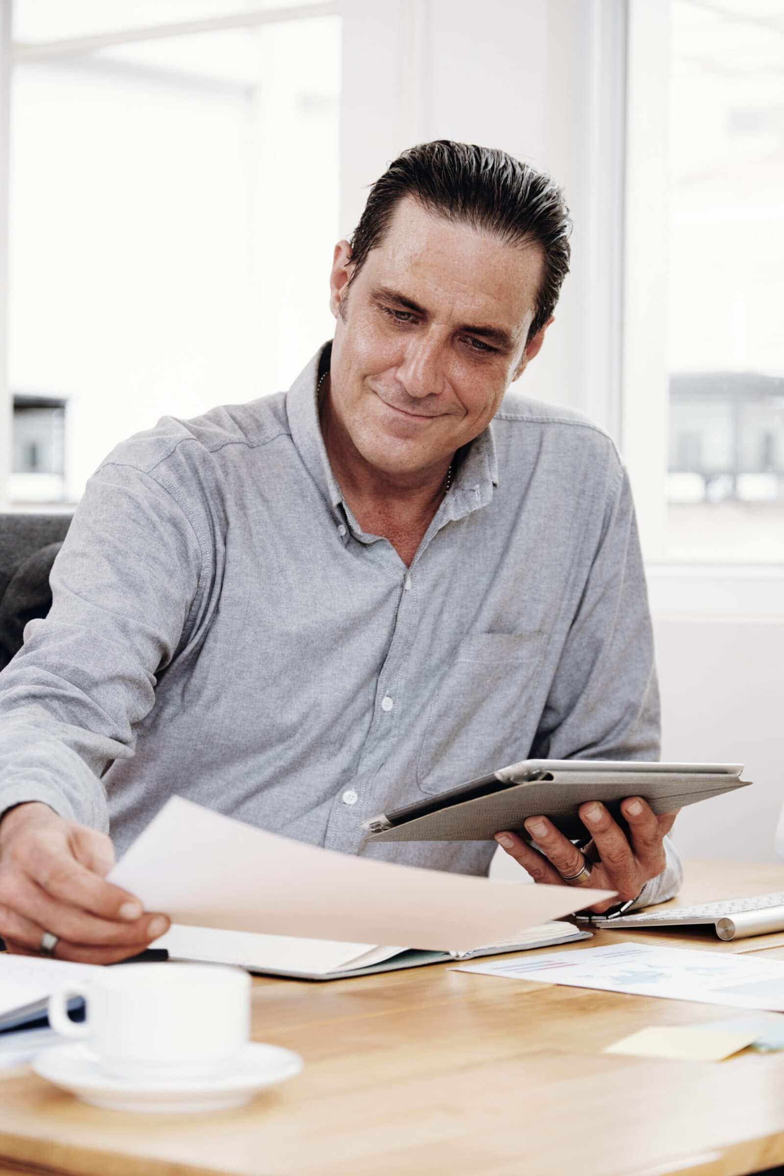 Um homem sentado em uma mesa, ocupado com um tablet e papéis espalhados, ilustrando um ambiente de trabalho produtivo